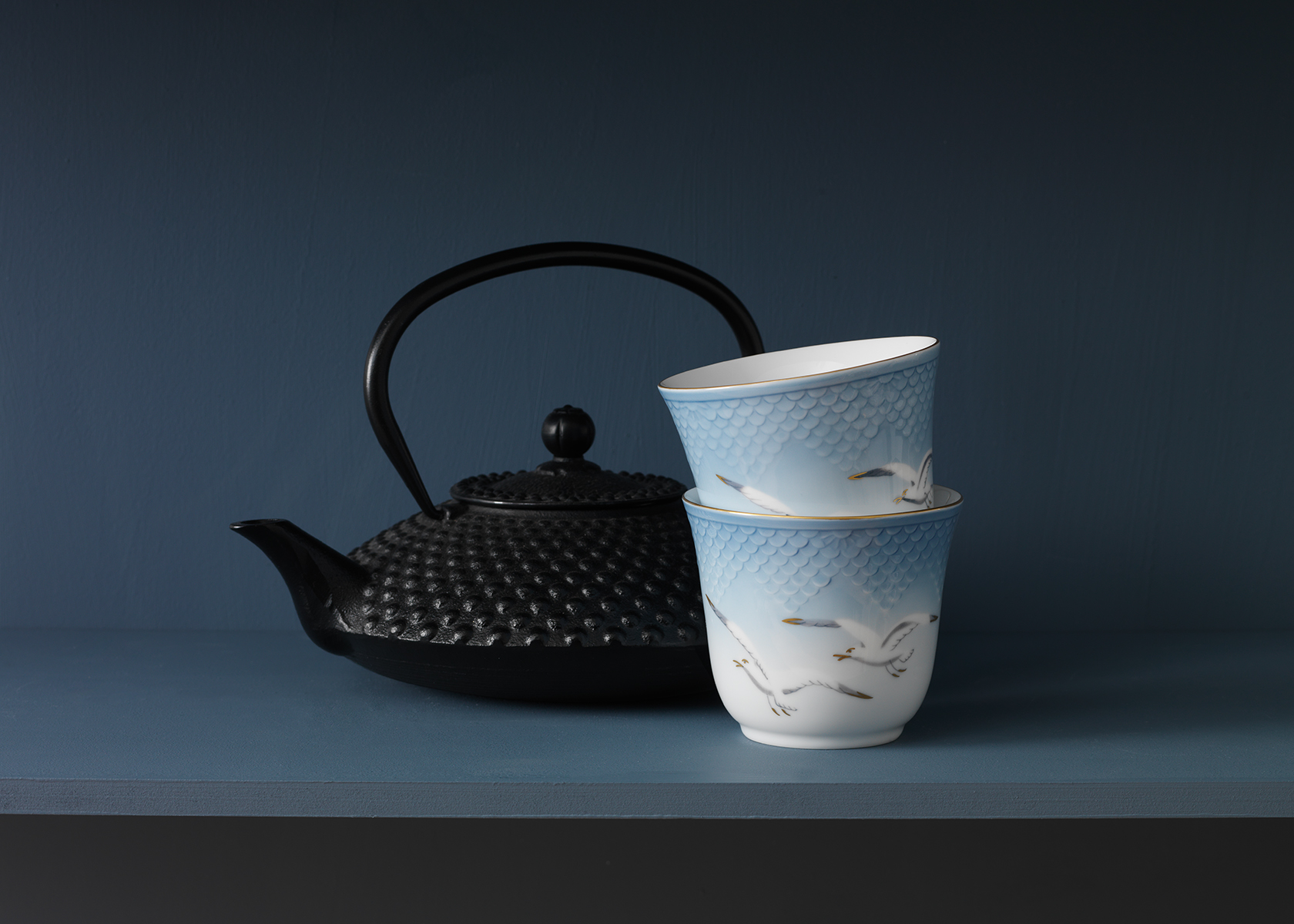 Two Seagull Thermal mugs stacked on a blue table in front of a black Teapot